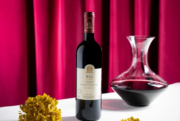 Flasche BEL Syrah Rotwein aus Malta neben einer Karaffe vor rotem Vorhang