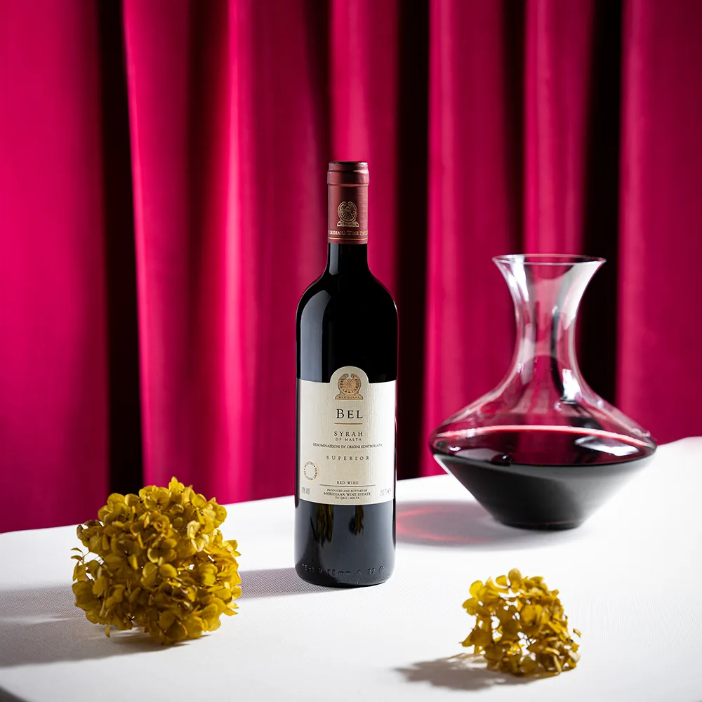Flasche BEL Syrah Rotwein aus Malta neben einer Karaffe vor rotem Vorhang