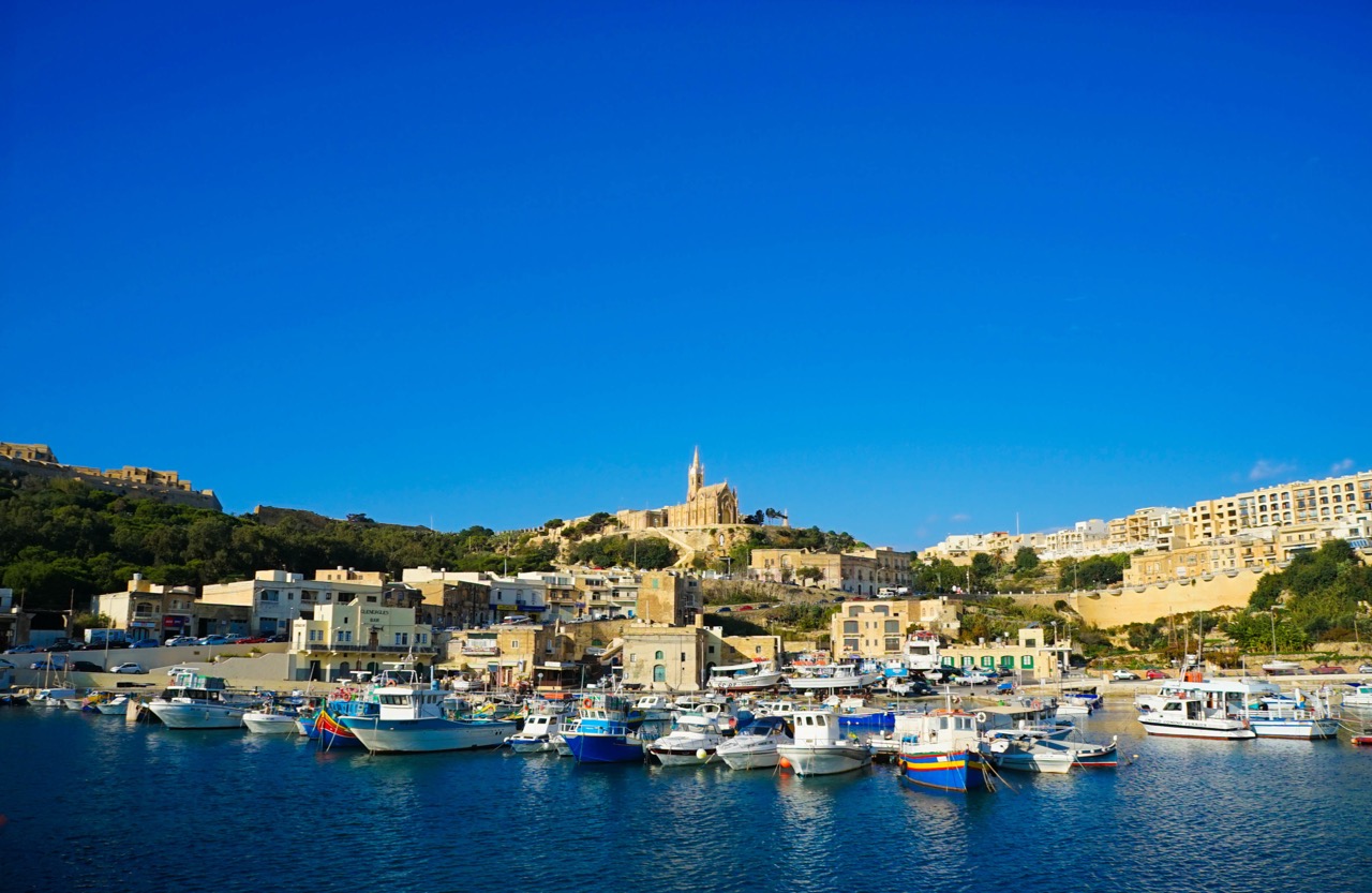 Hafen von Mġarr auf Gozo mit bunten Fischerbooten und Kirche am Hang