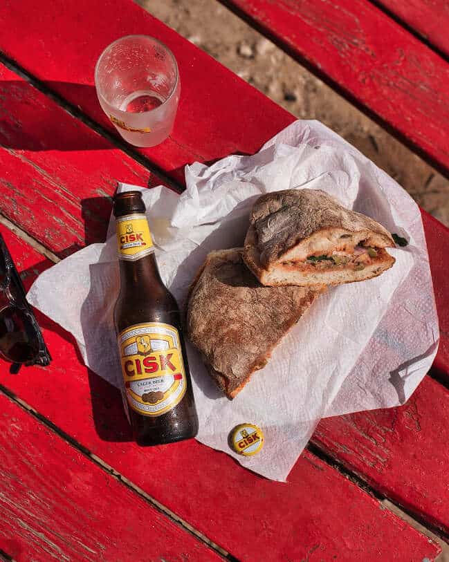 Cisk Lager Flasche und Glas neben einer maltesischen Ftira auf rotem Holztisch