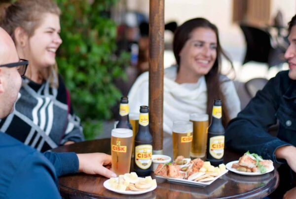 Freunde stoßen mit Cisk Lager Flaschen und Gläsern an einem Bartisch auf Malta an