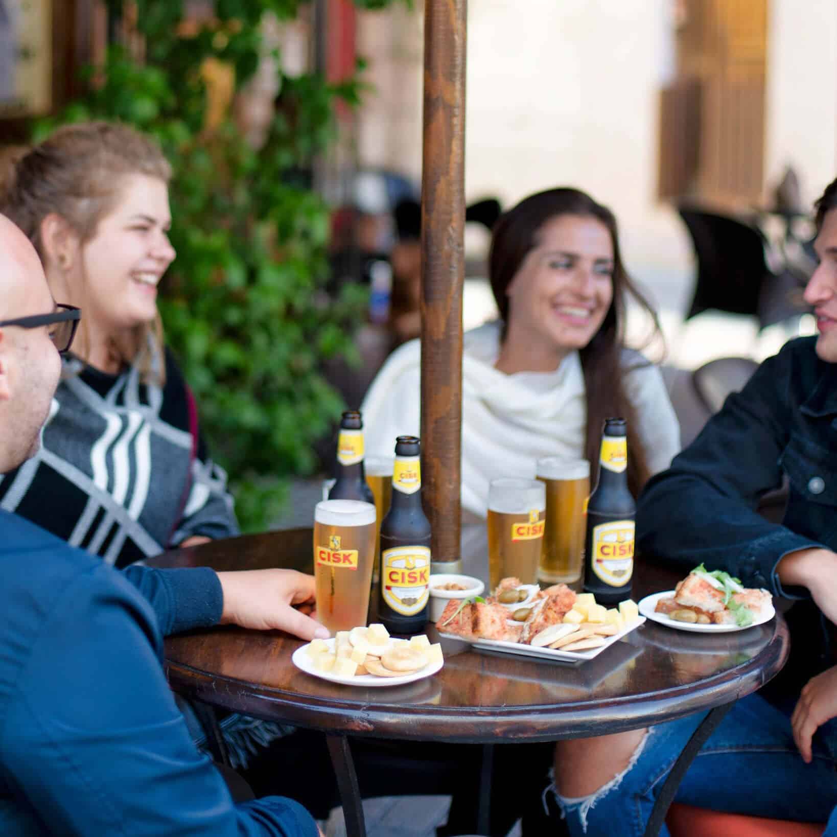 Freunde stoßen mit Cisk Lager Flaschen und Gläsern an einem Bartisch auf Malta an