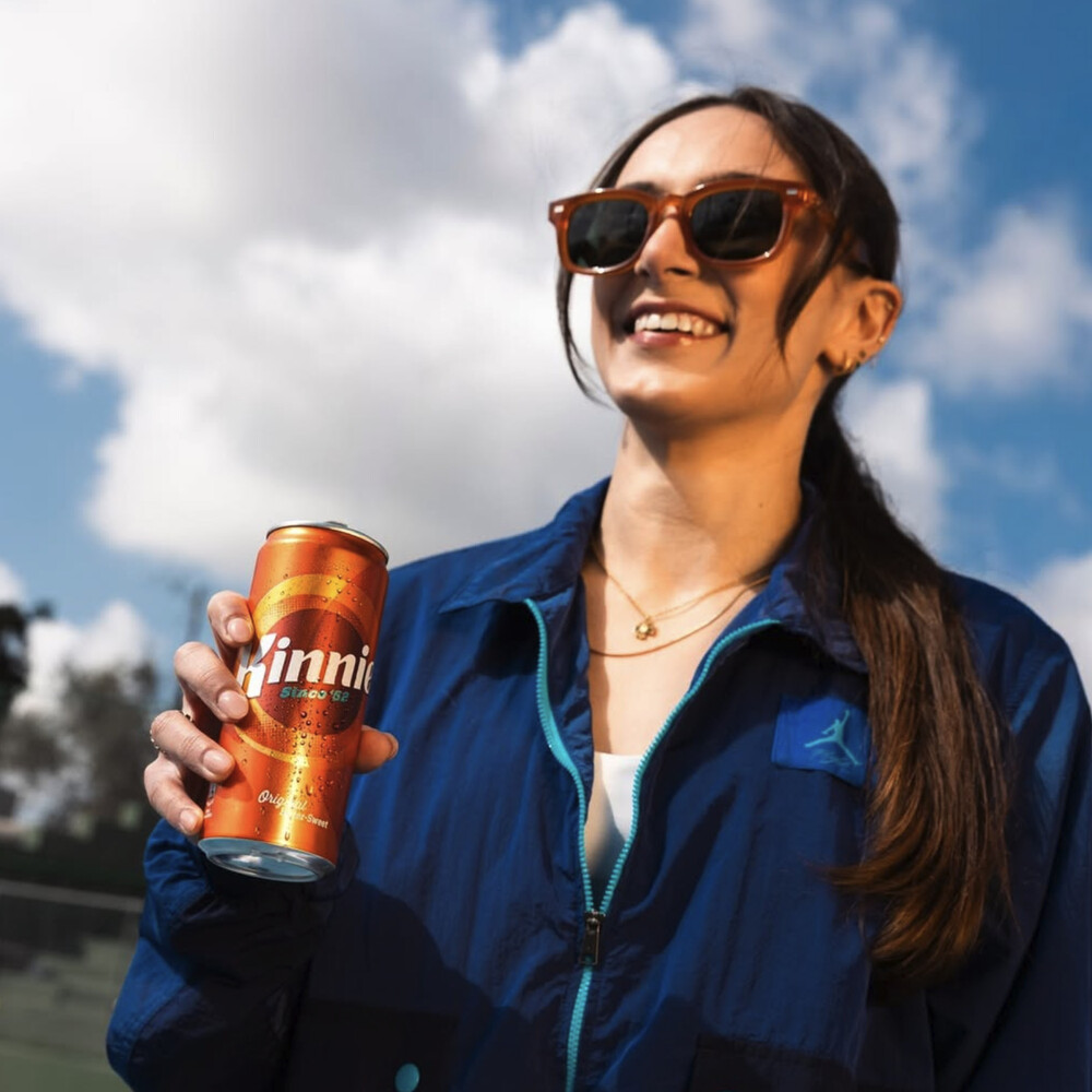 Junge Frau mit Sonnenbrille hält eine Kinnie Classic 330-ml-Dose in der Sonne